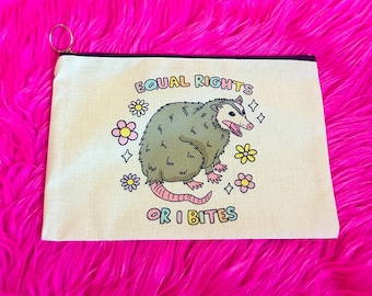 May include: Cream-colored zippered pouch featuring a cartoon opossum with the text "EQUAL RIGHTS OR I BITES." The opossum is green with a pink tail and is surrounded by colorful flowers and stars. The pouch has a gold-colored ring attached.