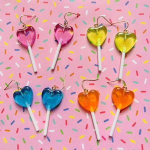 Op de afbeelding: Zes paar hartvormige lolly-oorbellen. De lolly's zijn in roze, geel, blauw en oranje. De oorbellen zijn gemaakt van hars en hebben gouden haakjes.