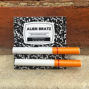 May include: Two white and orange cigarette-shaped hair clips with a red stripe. The clips are on a notebook paper background with a business card that says "Alien Bratz" with the website www.alienbratz.com, Instagram @Shop.alienbratz, and TikTok: Alien Bratz.