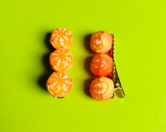 May include: Two orange hair clips with a design of orange slices. Each clip has four orange slices and a gold metal clip.