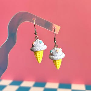 Op de afbeelding: Een paar hangende oorbellen met miniatuur ijshoorntjes met getextureerde lichtpaarse bollen en felgele wafelhoorntjes. Elke bol is versierd met kleine, kleurrijke stervormige spikkels en hangt aan een zilverkleurige metalen haak.