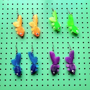 Pode incluir: Oito pares de brincos coloridos em forma de peixe. Os brincos são feitos de plástico e têm um acabamento brilhante. As cores incluem laranja, verde, azul e roxo.