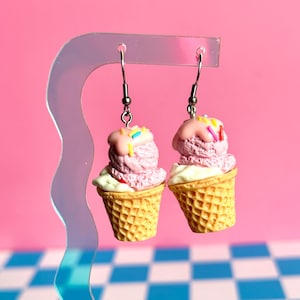 Op de afbeelding: Een paar hangende oorbellen met miniatuur ijshoorntjes met een gewafeld textuurpatroon, lichtpaarse ijsbolletjes, roze glazuur en kleurrijke spikkels, bevestigd aan zilverkleurige metalen oorhaken.