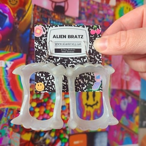 Pode incluir: Um par de brincos brancos translúcidos em forma de dentes. Os brincos estão pendurados em ganchos prateados. Os brincos estão em um cartão com o texto "ALIEN BRATZ" e o endereço do site "www.alienbratz.com".