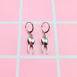 Pode incluir: Um par de brincos de prata em forma de dente com fecho de argola. Os brincos estão sobre um fundo rosa.