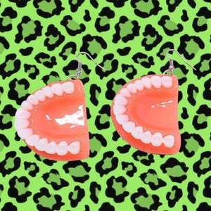 Pode incluir: Um par de brincos de plástico rosa e branco em forma de dentadura. Os brincos estão pendurados em ganchos prateados.