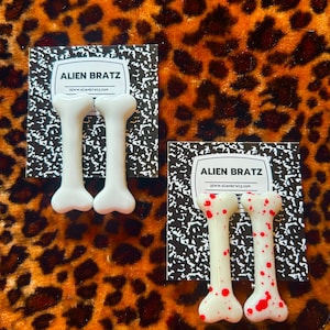 Pode incluir: Dois pares de brincos em forma de osso. Um par é branco, o outro é branco com respingos vermelhos. Ambos os pares estão em um fundo preto e branco com o texto "ALIEN BRATZ" e "www.alienbratz.com".