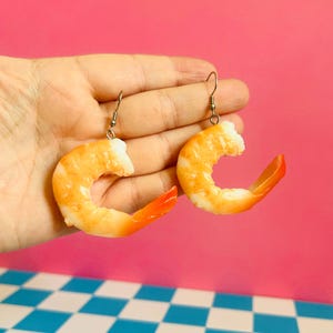 Op de afbeelding: Een paar hangende oorbellen in de vorm van gekookte garnalen. Elke oorbel heeft een realistisch, gebogen, oranje en wit garnalenlichaam dat aan een dunne zilverkleurige metalen haak hangt. De garnalen hebben een glanzende C-vorm.