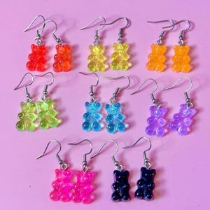 Pode incluir: Um conjunto de 10 pares de brincos de ursinho de goma em várias cores, incluindo vermelho, laranja, amarelo, verde, azul, roxo, rosa e preto. Os brincos são feitos de plástico transparente e têm a forma de um ursinho de goma.
