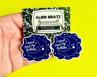 May include: A pair of dark blue earrings with a crescent moon and star design. The earrings have the text "I read past my bedtime". The earrings are on a white and black patterned card with the text "Alien Bratz".