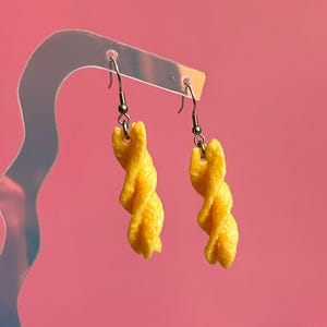 Op de afbeelding: Een paar bungelende oorbellen met gele, getextureerde, spiraalvormige pasta-bedels. Elke bedel hangt aan een zilverkleurige metalen oorhaakje. De oorbellen worden gepresenteerd op een doorzichtige, golvende acrylstandaard.