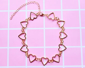 Op de afbeelding: Goudkleurige choker ketting met open hartvormige schakels. De ketting heeft een verstelbare ketting. Het ontwerp is eenvoudig en elegant, geschikt voor diverse gelegenheden. De ketting wordt gepresenteerd op een roze geruit achtergrond.