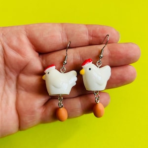 Pode incluir: Um par de brincos de novidade com figuras de galinhas brancas com chapéus vermelhos e bicos amarelos. Cada galinha pende de um gancho prateado e tem um ovo laranja pendurado. O fundo é amarelo brilhante.