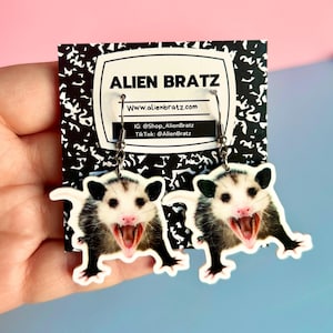 Op de afbeelding: Een paar zilveren creolen met een afbeelding van een wit en zwart opossum op elke oorbel. De oorbellen bevinden zich op een kaart met de tekst "Alien Bratz" en het websiteadres "www.alienbratz.com".