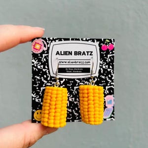 May include: A pair of corn on the cob earrings with silver hooks. The earrings are on a white card with the text "ALIEN BRATZ www.alienbratz.com IG: Shop_Alienbratz Tiktok: Alienbratz".