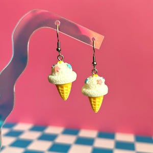 Op de afbeelding: Een paar hangende oorbellen met miniatuur ijsje-bedels. Elke bedel heeft een textuurrijke witte bol ijs met kleine, kleurrijke stervormige spikkels en een gele wafelhoorn, opgehangen aan zilverkleurige metalen haken.