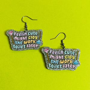 Puede incluir: Un par de pendientes novedosos con un borde brillante y el texto "Feelin cute might clog the work toilet later". Cada pendiente presenta un desatascador rosa y un gráfico de desatascador de inodoro azul.