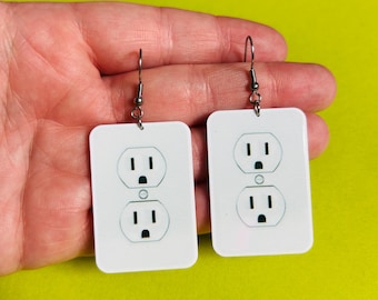 May include: A pair of novelty earrings featuring a white rectangular design with a black electrical outlet graphic. The earrings have silver-colored hooks and are displayed against a bright green background.