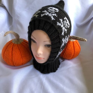 May include: Black and white knitted hat with a skull and crossbones pattern. The hat has a long neck and a pointed top. The hat is sitting on a white surface with two orange knitted pumpkins.