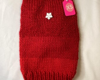 May include: A bright red, hand-knitted dog sweater with a ribbed collar and hem. Two white snowflake-shaped buttons adorn the front. A pink tag with "XS" is attached. The sweater is designed for a small dog.