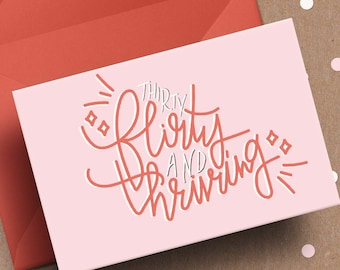 May include: A pink greeting card with the text "Thirty, Flirty, and Thriving" in coral script, next to a coral envelope. The background is a neutral brown with scattered pink and white confetti.