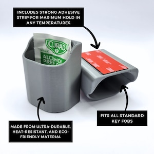 May include: A gray, 3D-printed key fob holder with a strong adhesive strip. The holder includes the text "INCLUDES STRONG ADHESIVE STRIP FOR MAXIMUM HOLD IN ANY TEMPERATURES" and "FITS ALL STANDARD KEY FOBS". It is made from ultra-durable, heat-resistant, and eco-friendly material.
