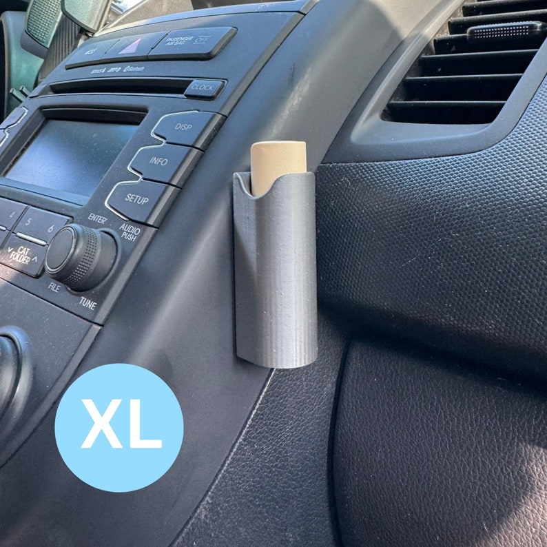 Op de afbeelding: Een grijze plastic autotelefoonhouder met een beige lippenbalsem erin. De houder is bevestigd aan de zijkant van het dashboard van een auto. De tekst "XL" is zichtbaar in de linkerbenedenhoek van de afbeelding.