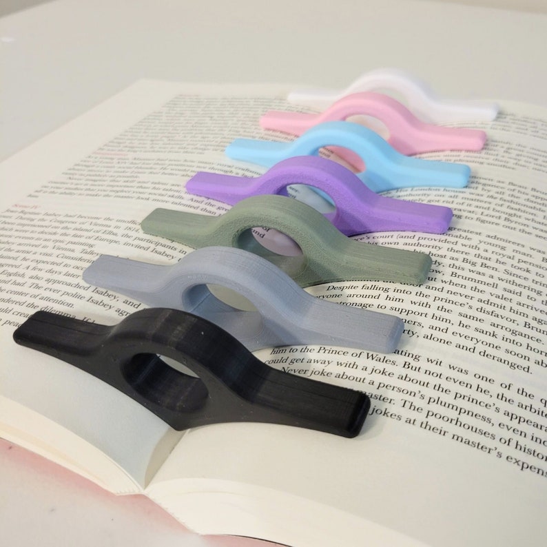 May include: A collection of colorful, curved book page holders in various colors, including black, gray, green, purple, blue, pink, and white. These reading accessories are designed to hold a book open. The book is open to a page of printed text.