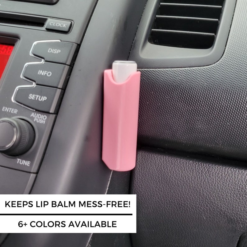 May include: A pink lip balm holder attached to the inside of a car. The holder has a white label with the text "KEEPS LIP BALM MESS-FREE! 6+ COLORS AVAILABLE".