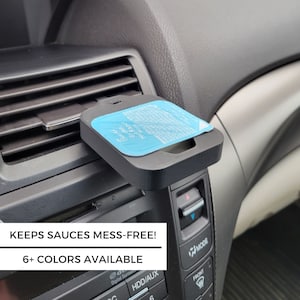 May include: A black plastic car air vent mount with a blue sauce packet holder. The text "Here Ranch" and "Garden Sauce" is visible on the sauce packet. The caption "Keeps Sauces Mess-Free!" and "6+ Colors Available" is visible below the image.