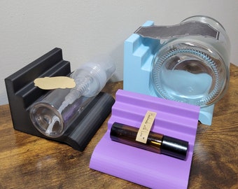 May include: Three different sized, black, purple, and light blue, 3D printed soap or lotion bottle holders. The holders are designed to hold bottles upright. The holders are on a wooden surface. The purple holder has a small brown label that says "Handmade with Love".