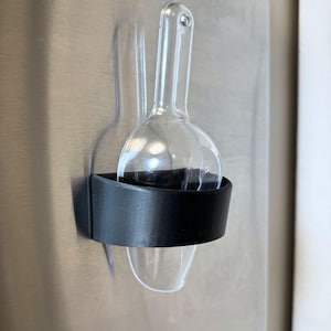 May include: A clear plastic ice cream scoop with a black plastic holder attached to a stainless steel surface.