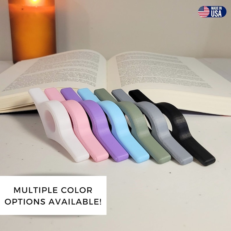 May include: A collection of colorful book holders in various shades, including white, pink, purple, and blue, arranged in a row. The text "MULTIPLE COLOR OPTIONS AVAILABLE!" is displayed below the book holders. The image also includes an open book and a lit candle.