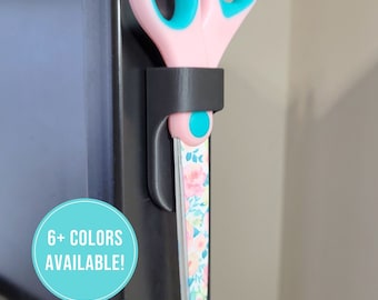 May include: A black plastic holder with a pink and turquoise pair of scissors with a floral pattern on the blades. The text "6+ COLORS AVAILABLE!" is on a turquoise circle.