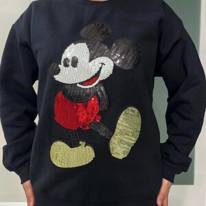 Op de afbeelding: Zwart sweatshirt met een groot paillettenontwerp van Mickey Mouse. Het stripfiguur heeft een wit gezicht, een rode broek en gouden schoenen. De pailletten geven een sprankelend effect aan het klassieke cartoonbeeld.