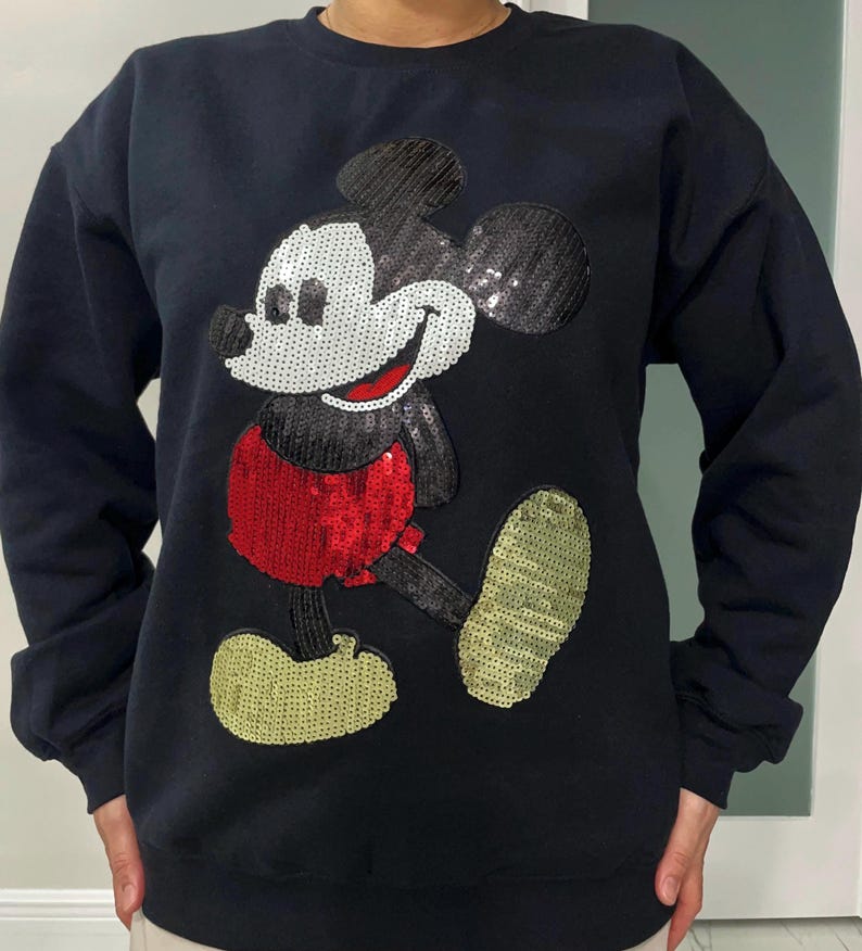Op de afbeelding: Zwart sweatshirt met een groot paillettenontwerp van Mickey Mouse. Het stripfiguur heeft een wit gezicht, een rode broek en gouden schoenen. De pailletten geven een sprankelend effect aan het klassieke cartoonbeeld.