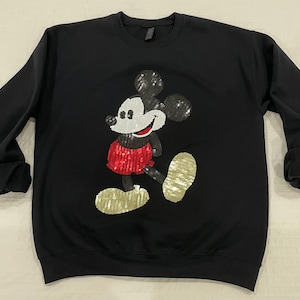 Op de afbeelding: Zwart sweatshirt met ronde hals met een pailletten Mickey Mouse-ontwerp. De Mickey Mouse-afbeelding heeft zwarte paillettenoren, een wit gezicht, een rode paillettenbroek en gouden paillettenschoenen. De sweatshirt heeft lange mouwen met opgerolde manchetten.