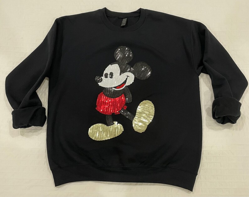 Op de afbeelding: Zwart sweatshirt met ronde hals met een pailletten Mickey Mouse-ontwerp. De Mickey Mouse-afbeelding heeft zwarte paillettenoren, een wit gezicht, een rode paillettenbroek en gouden paillettenschoenen. De sweatshirt heeft lange mouwen met opgerolde manchetten.