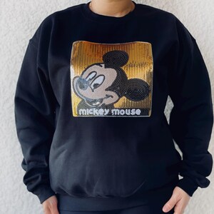 May include: Black sweatshirt featuring a sequined Mickey Mouse design. The design includes a gold sequined background with a Mickey Mouse graphic and the words "Mickey Mouse" in white.