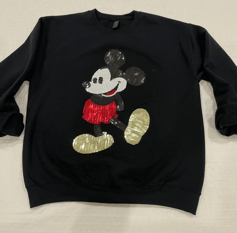 Op de afbeelding: Zwart sweatshirt met een pailletten Mickey Mouse-ontwerp. Het stripfiguur wordt wandelend getoond, met een rode paillettenrok en gouden paillettenschoenen. De sweatshirt heeft lange mouwen en een ronde hals.