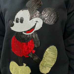 Op de afbeelding: Zwart sweatshirt met een Mickey Mouse-ontwerp van pailletten. Het stripfiguur wordt wandelend afgebeeld, met een wit gezicht, een rode broek en gele schoenen. De pailletten geven een glinsterend effect aan het klassieke stripfiguur.