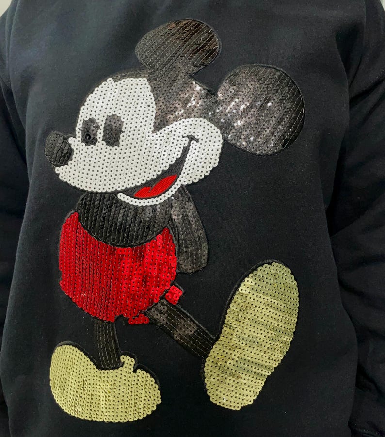 Op de afbeelding: Zwart sweatshirt met een Mickey Mouse-ontwerp van pailletten. Het stripfiguur wordt wandelend afgebeeld, met een wit gezicht, een rode broek en gele schoenen. De pailletten geven een glinsterend effect aan het klassieke stripfiguur.