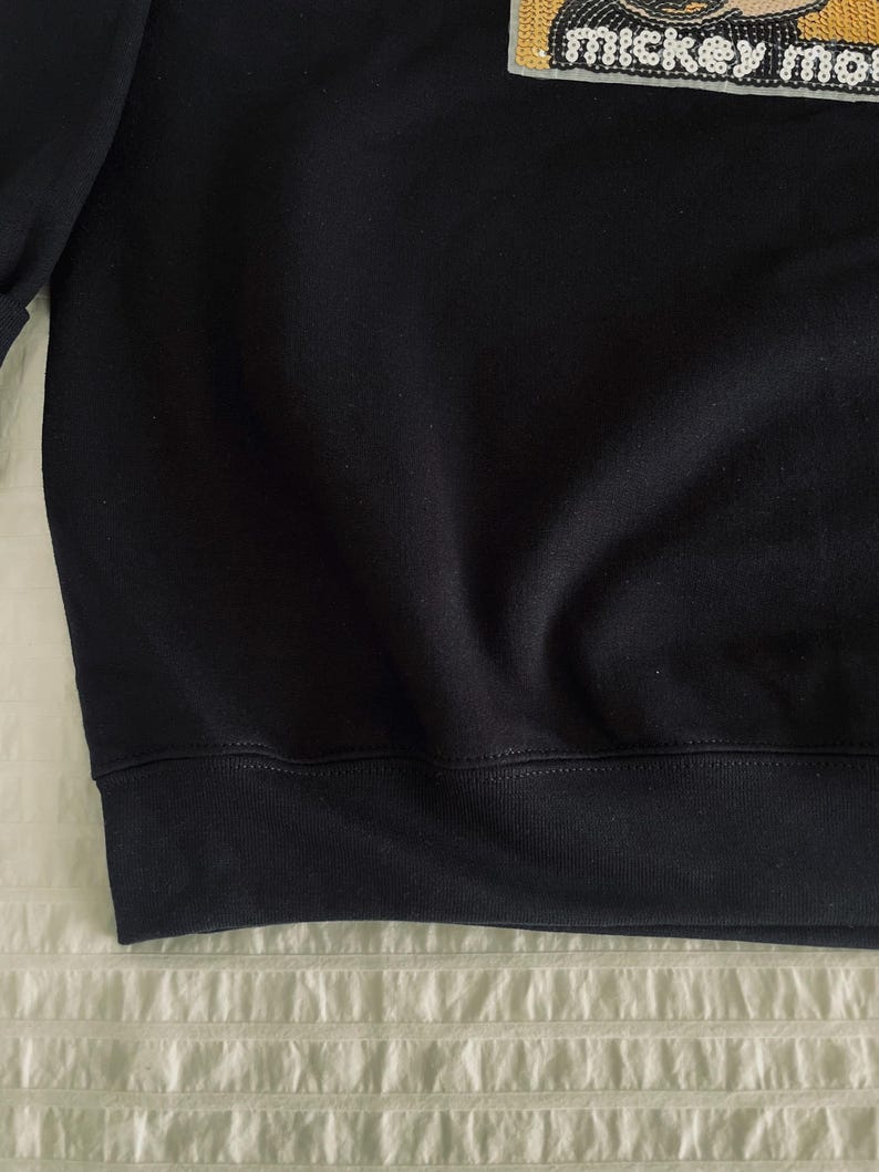 Op de afbeelding: Zwart sweatshirt met geribbelde zoom en manchetten. De rechterbovenhoek heeft een patch met de tekst "Mickey Mouse" in een decoratief lettertype. Het sweatshirt is gemaakt van een zachte, donkere stof.