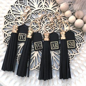 May include: Several black tassel keyrings with gold-coloured details and initialled tags. Each keyring has a gold clasp and a long, black tassel. The tags display the initials "TG", "VG", "AR", and "SM" in a gold font.