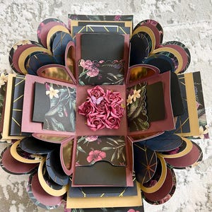 May include: A black and gold explosion box with a floral design. The box is decorated with pink roses, gold accents, and a black and white geometric pattern. The box is open and the inside is visible, revealing multiple layers of paper and a small compartment.
