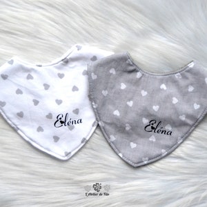 May include: Two baby bibs, one white and one gray, each patterned with small gray hearts. The name "Eléna" is printed in black script on each bib. The bibs are triangular in shape.