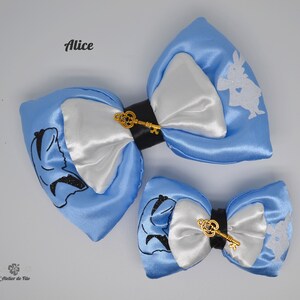 Puede incluir: Dos lazos para el pelo de satén azul claro con centros de satén blanco y detalles en negro. Un lazo presenta la silueta de Alicia, el otro un conejo blanco. Ambos tienen dijes de llave dorados. La palabra "Alice" está impresa en la parte superior izquierda.