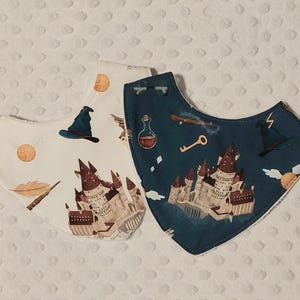 May include: Two baby bibs with a fantasy theme. One bib is white with a castle, a wizard hat, and other graphics. The other bib is dark blue with a castle, a potion bottle, and a key. Both bibs have a soft, white backing.