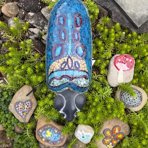 Könnte beinhalten: Eine blaue Mosaik-Insektenskulptur mit Gesichtsdesign, umgeben von bemalten dekorativen Steinen. Der Käfer hat rote und violette Akzente. Die Steine weisen verschiedene Designs auf, darunter ein Herz, eine Blume und ein Pilz.
