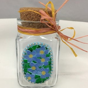 Pode incluir: Um frasco de vidro transparente com tampa de cortiça e um mosaico azul e verde de flores de miosotis. O frasco é decorado com uma fita amarela e rosa.